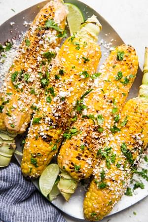 Elote (Mexican street corn) on a platter, topped with chipotle mayo, cotija cheese, and cilantro, served with lime wedges.