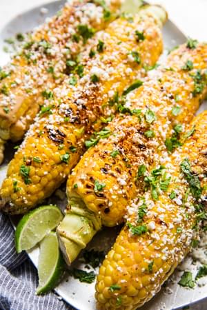 Elote (Mexican street corn) on a platter, topped with chipotle mayo, cotija cheese, and cilantro, served with lime wedges.