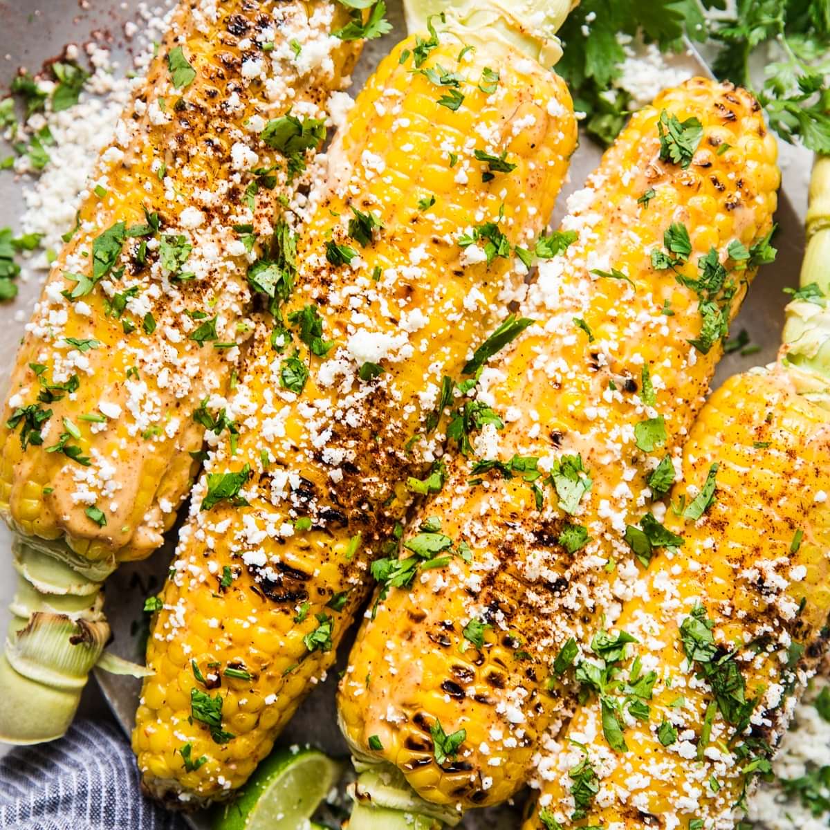 Elote (Mexican street corn) on a platter, topped with chipotle mayo, cotija cheese, and cilantro, served with lime wedges.
