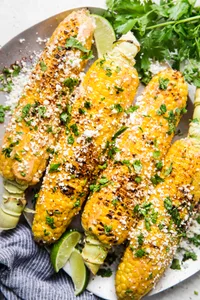 Elote (Mexican street corn) on a platter, topped with chipotle mayo, cotija cheese, and cilantro, served with lime wedges.
