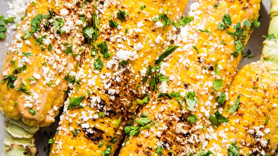 Elote (Mexican street corn) on a platter, topped with chipotle mayo, cotija cheese, and cilantro, served with lime wedges.