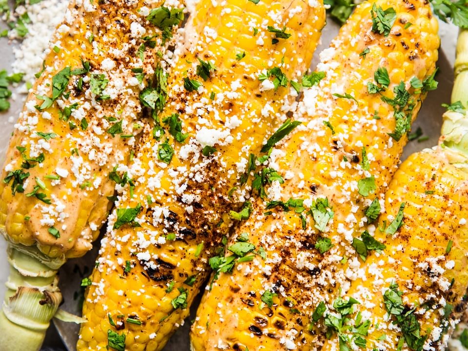 Elote (Mexican street corn) on a platter, topped with chipotle mayo, cotija cheese, and cilantro, served with lime wedges.