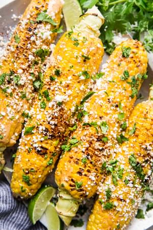 Elote (Mexican street corn) on a platter, topped with chipotle mayo, cotija cheese, and cilantro, served with lime wedges.