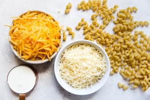 grated cheddar cheese, parmesan cheese, whole milk and raw pasta in bowls to make stovetop mac and cheese