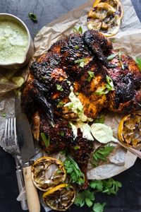 whole grilled chicken spatchcocked on parchment paper with a bowl of green garlic sauce grilled lemons