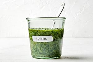 a jar of basil pesto with a spoon in it made with basil, Parmesan, pine nuts, cloves, sea salt and olive oil