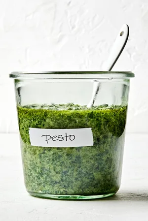 a jar of basil pesto with a spoon in it made with basil, Parmesan, pine nuts, cloves, sea salt and olive oil