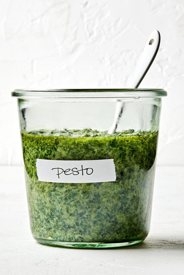 a jar of basil pesto with a spoon in it made with basil, Parmesan, pine nuts, cloves, sea salt and olive oil