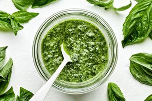 a jar of basil pesto with a spoon in it made with basil, Parmesan, pine nuts, cloves, sea salt and olive oil