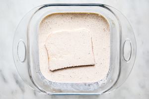 a slice of brioche bread being dipped cream and egg batter in a glass dish on the counter for homemade French toast