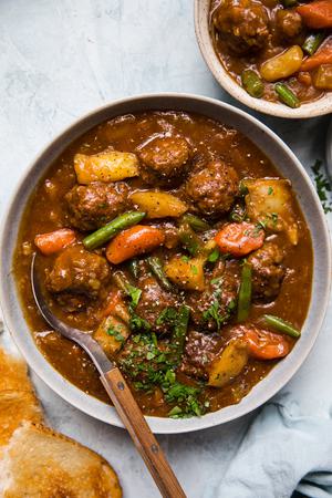 a bowl of homemade meatball stew with a spoon topped with parsley and a pice of crunchy bread beside it