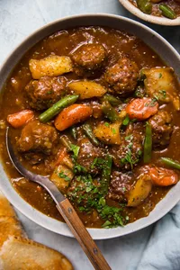 a bowl of homemade meatball stew with a spoon topped with parsley and a pice of bread beside it