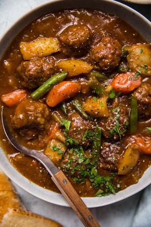 a bowl of homemade meatball stew with a spoon topped with parsley and a pice of crunchy bread beside it