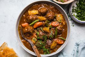 a bowl of homemade meatball stew with a spoon topped with parsley and a pice of crunchy bread beside it