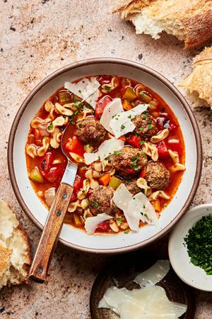 a bowl of meatball soup topped with parmesan and parsley surrounded by crusty bread and bowls of extra parmesan and parsley