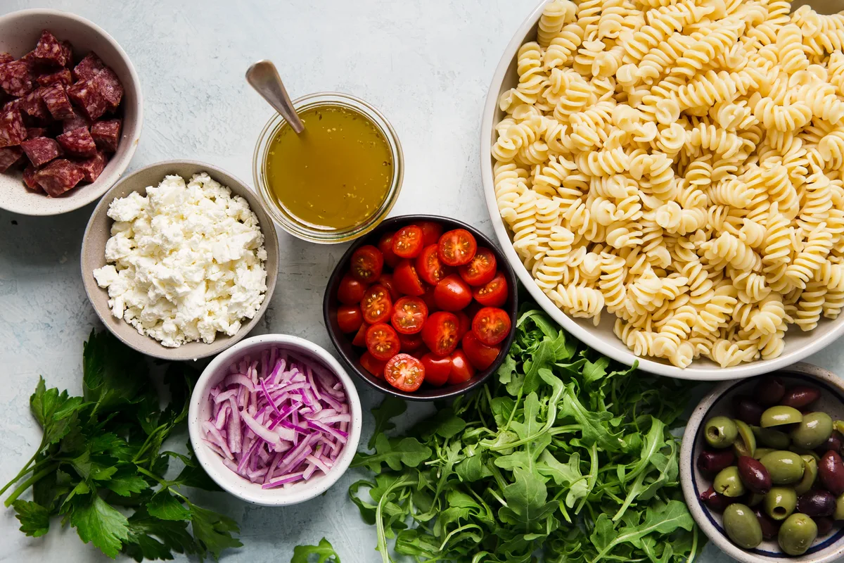 ingredients laid out pasta, olives, arugula, tomatoes, red onions, feta and salami