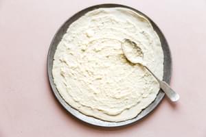 spreading white garlic sauce on pizza crust with a spoon