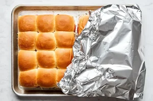 Sliced Hawaiian rolls filled with pizza sauce, cheese and pepperoni on a baking sheet being covered with foil