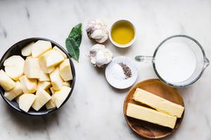 a bowl of peeled chopped potatoes, 2 bay leaves, whole milk, butter, 2 garlic heads, salt and pepper in bowls on a counter