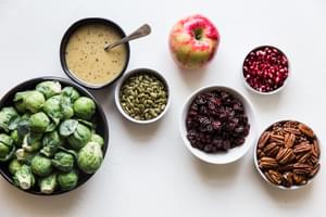 Brussels sprouts in a bowl and bowls filled with maple dressing, pipits, pecans, pomegranates, dried cherries and an apple