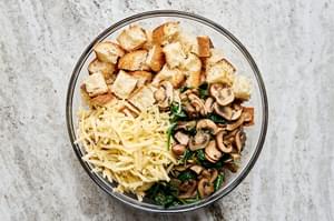 toasted cubed bread, cooked spinach, cooked mushrooms, cooked onion and Swiss cheese being tossed in a bowl