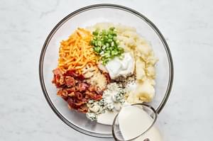 potato, bacon, cheddar, blue cheese, sour cream, cream, green onions, and spices being mixed together in a bowl