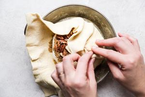 hand wrapping puffed pastry around wheel of brie topped with pecans, pepper and brown sugar in a baking dish