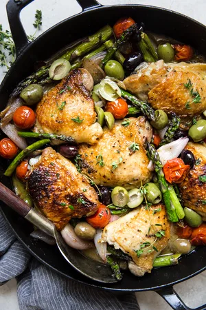 Baked chicken thighs in a cast iron skillet surrounded by asparagus, olives, and tomatoes.