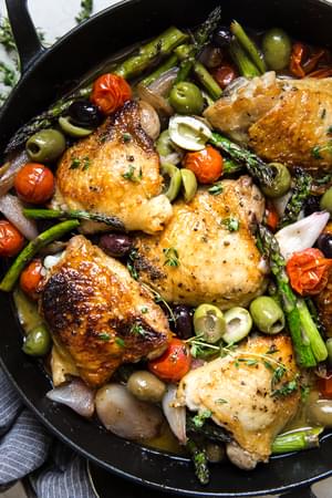 Baked Chicken Thighs With Asparagus in a pan with olives, tomatoes and shallots in a white wine garlic sauce.