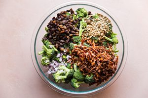 large bowl of raw broccoli, bacon, sunflower seeds, purple onions and golden raisins ready to be mixed with creamy dressing