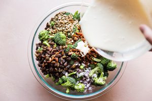 sweet creamy dressing being poured onto homemade broccoli salad with crispy bacon, golden raisins and purple onions