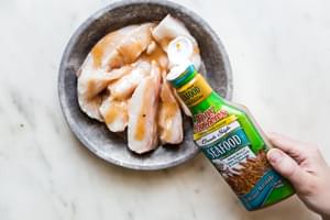 hand pouring tony's seafood marinade over white fish in a baking dish