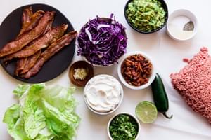 Ingredients laid out bacon, red cabbage, ground beef guacamole, lettuce, jalapeño, garlic.