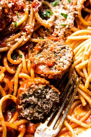 an Italian meatball cut in half with a fork on top of spaghetti and homemade sauce topped with parmesan and parsley