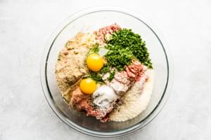 A large mixing bowl filled with fresh meatball ingredients: beef, pork, eggs, breadcrumbs, parsley, Parmesan and spices