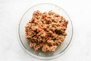 A large mixing bowl filled with fresh meatball ingredients: beef, pork, eggs, breadcrumbs, parsley, Parmesan and spices