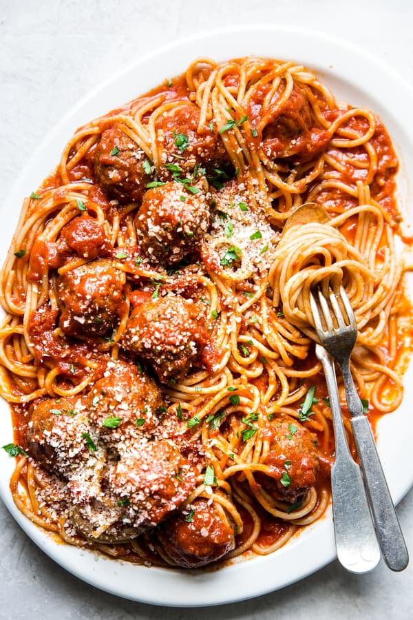 A platter of homemade spaghetti and meatballs topped with freshly grated Parmesan and parsley with a serving fork and spoon