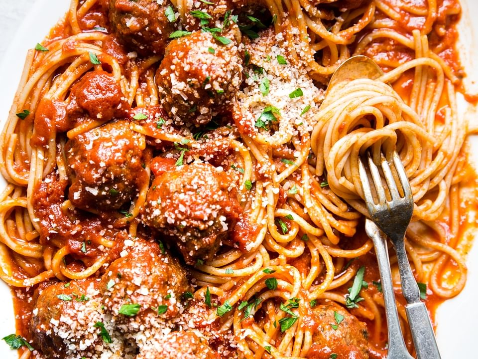 A platter of homemade spaghetti and meatballs topped with freshly grated Parmesan and parsley with a serving fork and spoon