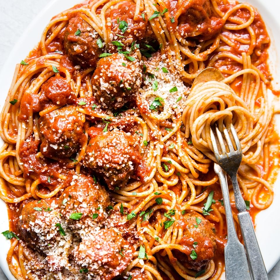 A platter of homemade spaghetti and meatballs topped with freshly grated Parmesan and parsley with a serving fork and spoon