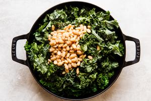 black cast iron skillet with leafy kale and white beans over crispy chicken thighs