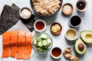 Ingredients laid out for a sushi salmon bowl avocado, cucumber, nori, cooked brown rice, sriracha, mayonnaise and soy sauce
