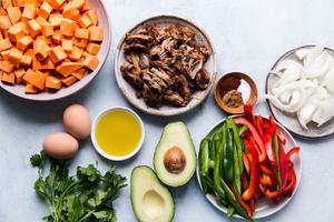Ingredients laid out: sweet potatoes, carnitas, avocado, olive oil, eggs, cilantro red and green bell peppers and onions.