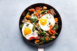 Sweet Potato Carnitas Hash in a pan with red and green bell peppers 2 fried eggs and cilantro