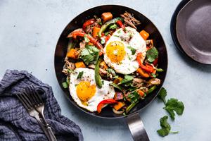 Sweet Potato Carnitas breakfast Hash in a pan with red and green bell peppers 2 fried eggs and cilantro with plates and forks
