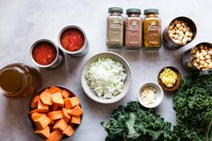 ingredients laid out 2 cans tomatoes spices, chickpeas, sweet potatoes, kale broth and onions.