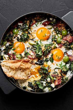 Baked eggs with swiss chard and green olives in a baking dish served with bread in a list of healthy breakfast recipes