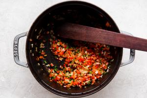 wooden spoon stirring jalapeño red bell peppers and onions added to rendered bacon fat in a soup pot.