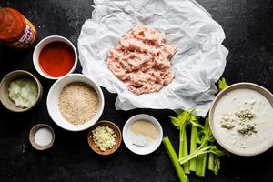 Ground chicken on parchment paper, a small bowl of but cheese dressing, celery, and buffalo sauce and meatball ingredients
