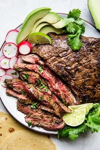 A plate of grilled carne asada with tortillas, limes, avocado and radishes