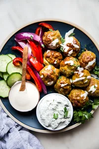 chicken shawarma meatballs on a blue plate shown with roasted red peppers, onions, cucumbers, tahini and tzatziki sauce.